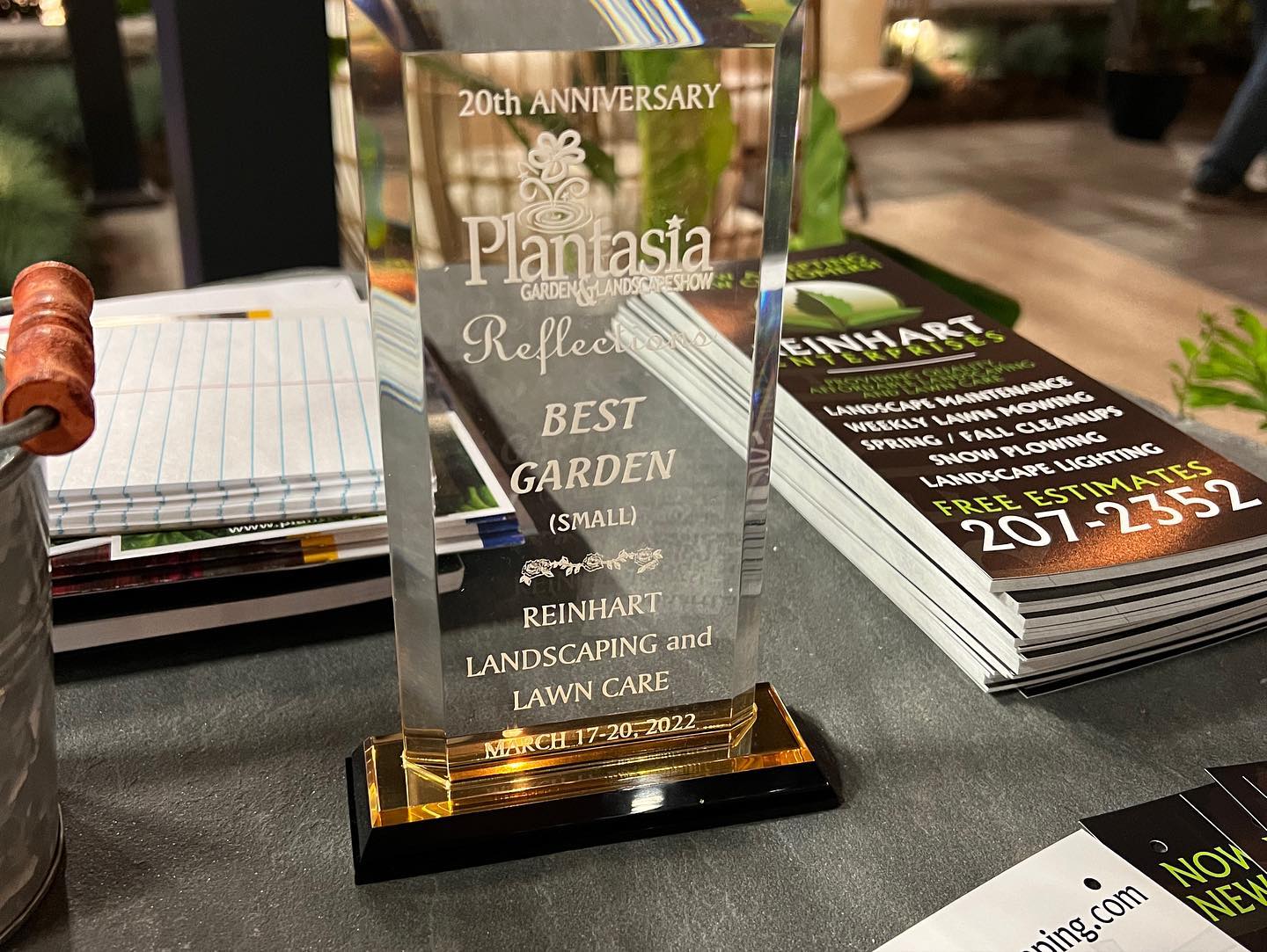 Clear glass award trophy engraved with “20th Anniversary Plantasia Reflections Best Garden (Small) Reinhart Landscaping and Lawn Care” displayed on a table next to stacks of Reinhart Enterprises Landscaping brochures and papers.